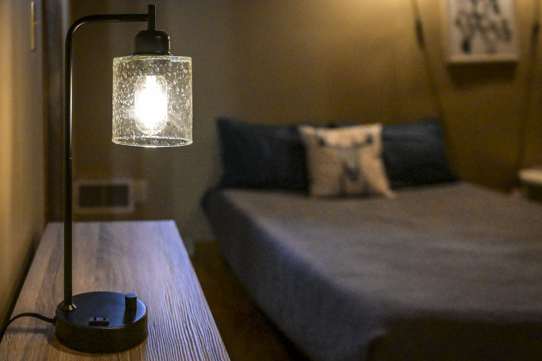 Et natbord med en lampe, en seng med blåt sengetæppe og puder, samt et billede hængt på væggen.