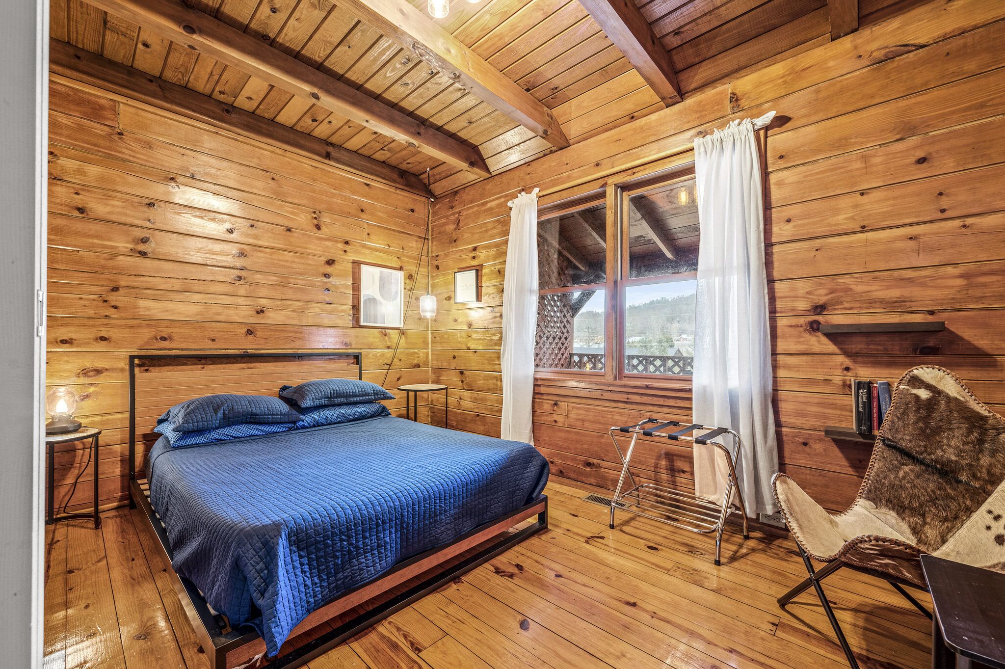 Habitació de cabana de fusta amb un llit, una cadira i una finestra amb cortines.