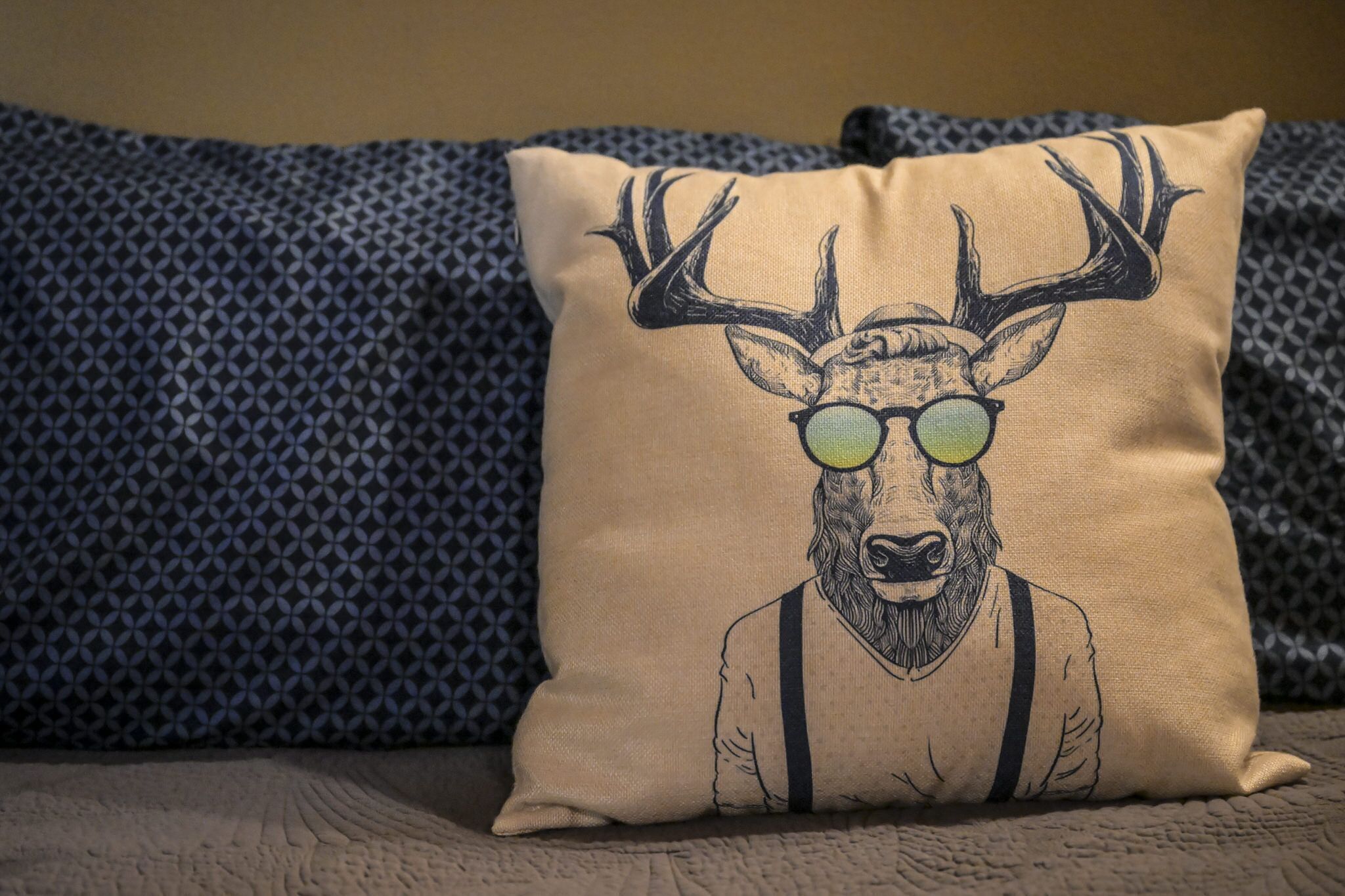 A beige pillow with a deer illustration on a textured blue and beige patterned bedspread.