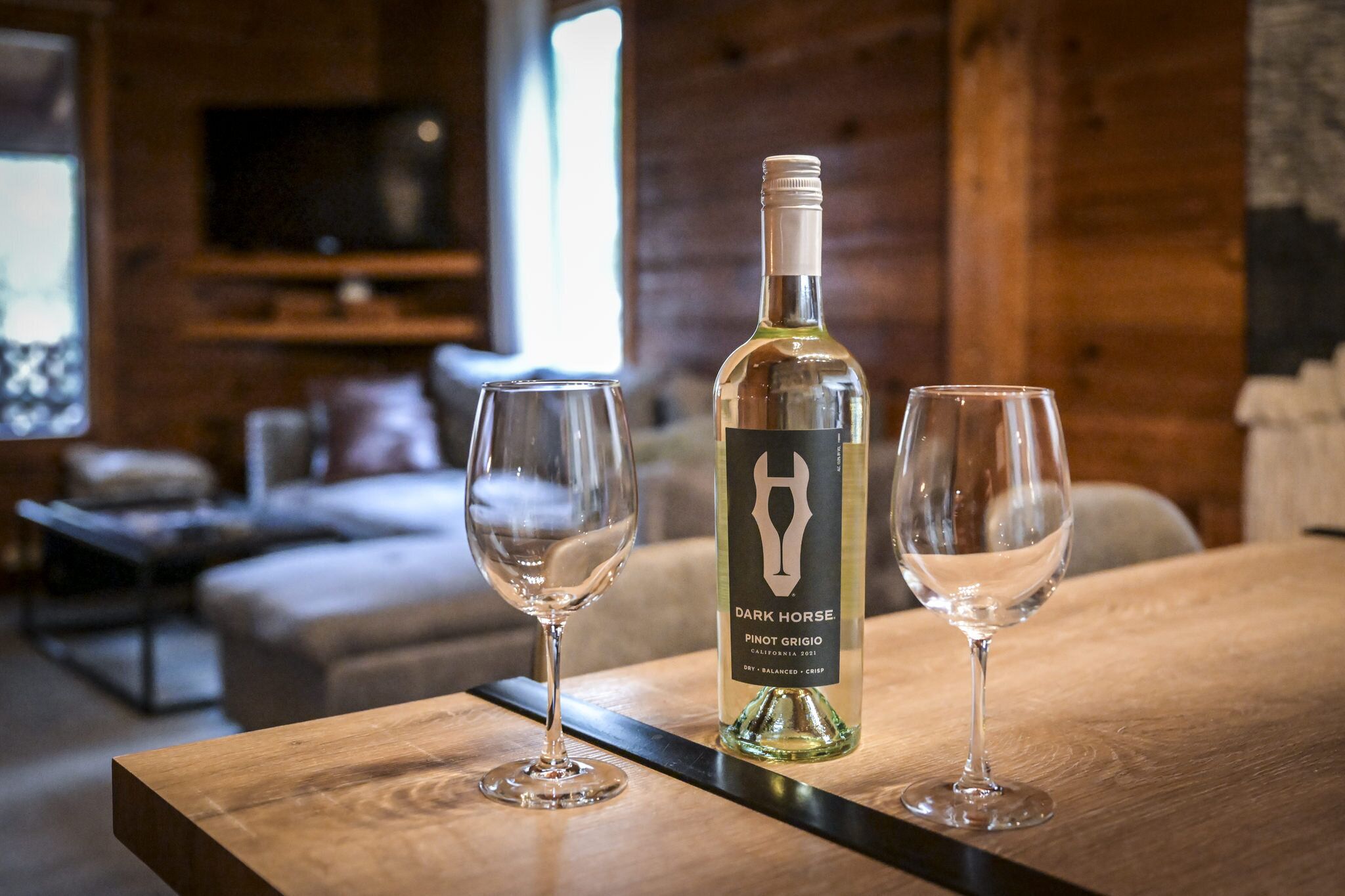 Une bouteille de vin Pinot Grigio de la marque Dark Horse et deux verres à vin vides sont posés sur une table en bois, dans une pièce chaleureuse au décor intérieur en bois, avec un téléviseur en arrière-plan.