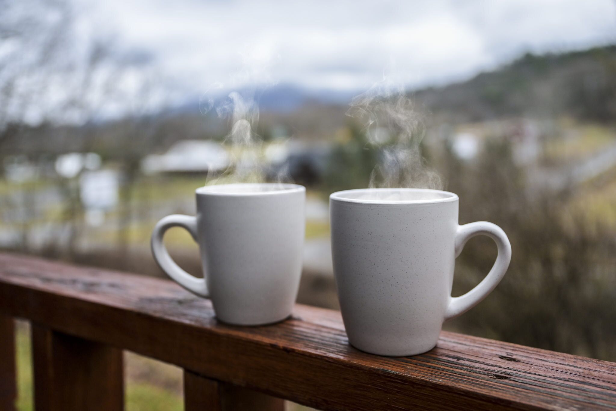 Two white ceramic mugs on a wooden railing with steam rising from them, set against a blurred background of a natural landscape.