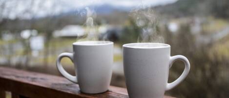 Two white ceramic mugs on a wooden railing with steam rising from them, set against a blurred background of a natural landscape.
