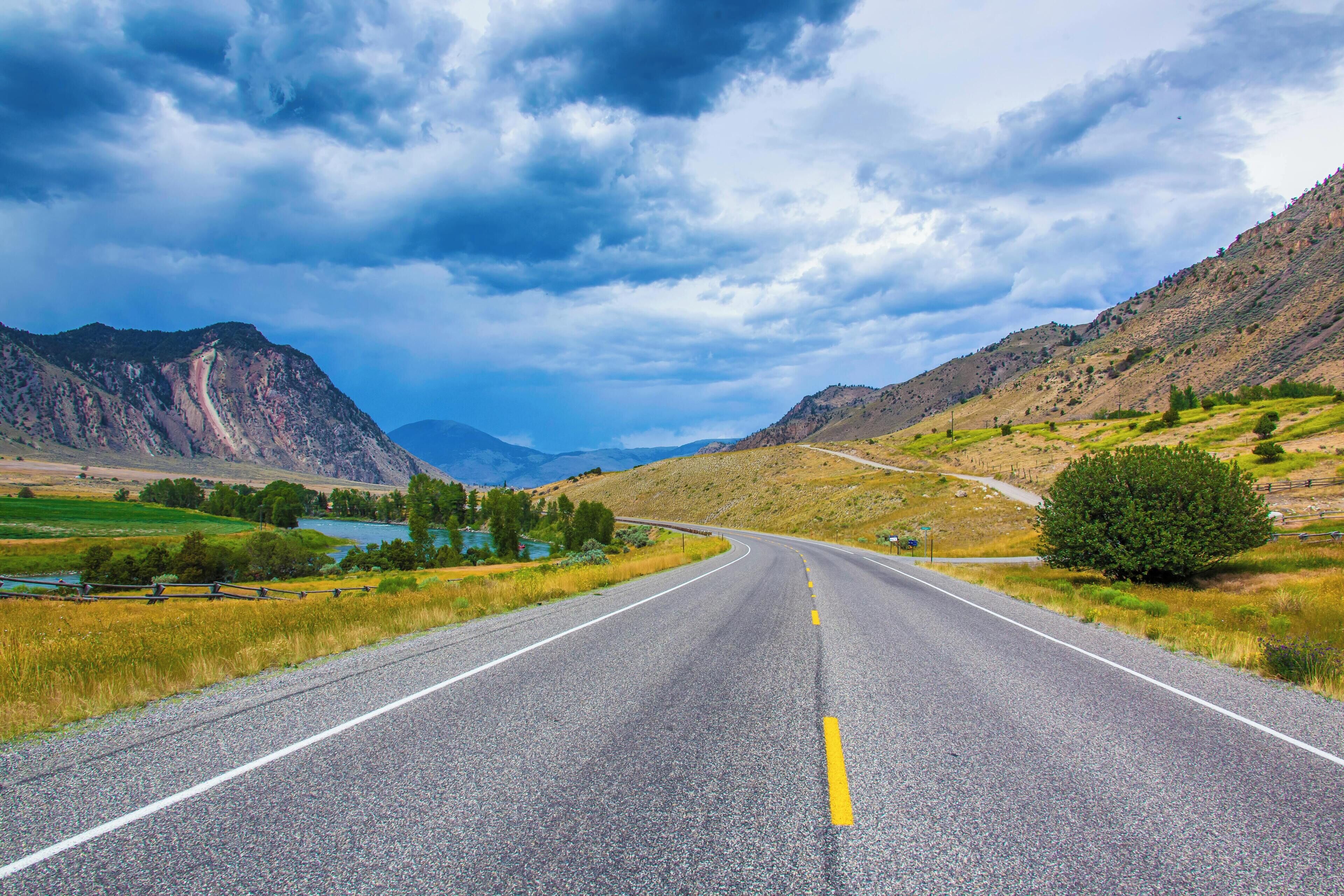 A straight road with a yellow center line, flanked by grassy fields and mountains under a cloudy sky.