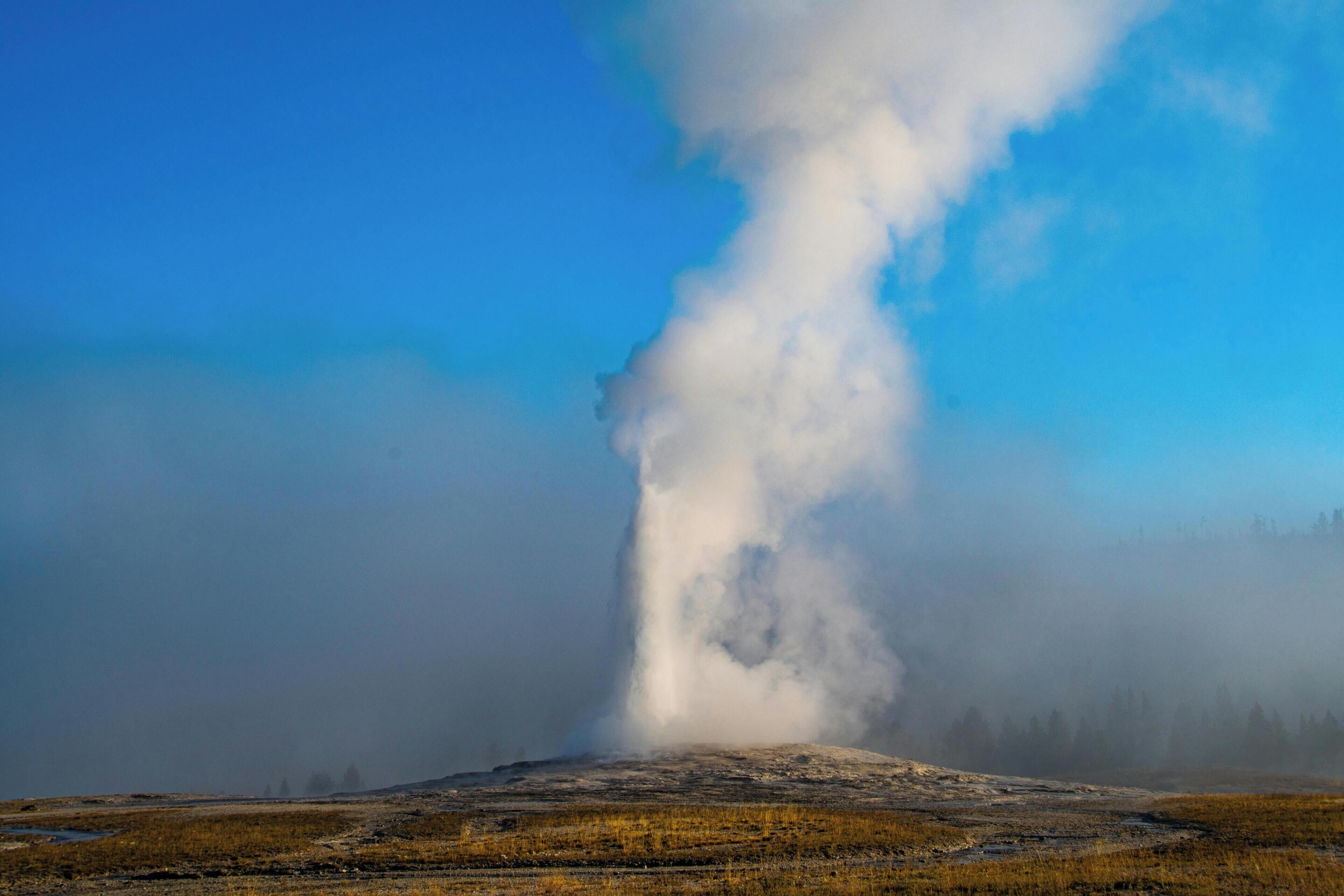 Ein Geysir mit aufsteigendem Dampf, umgeben von einer flachen Landschaft mit spärlicher Vegetation.