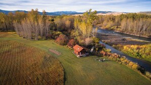 Une cabane rustique située à la campagne, près d’une rivière, et entourée de champs et d’arbres.