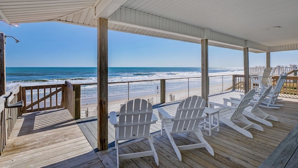 A wooden deck with white Adirondack chairs overlooking a beach.