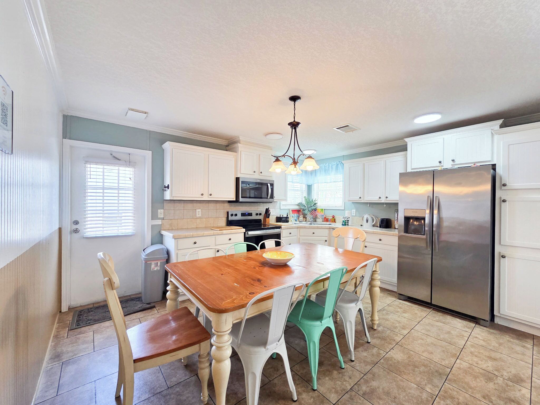 A kitchen with white cabinets, a stainless steel refrigerator, a wooden dining table with chairs, and a chandelier.