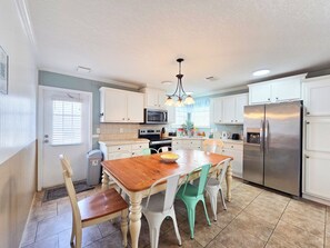A kitchen with white cabinets, a stainless steel refrigerator, a wooden dining table with chairs, and a chandelier.
