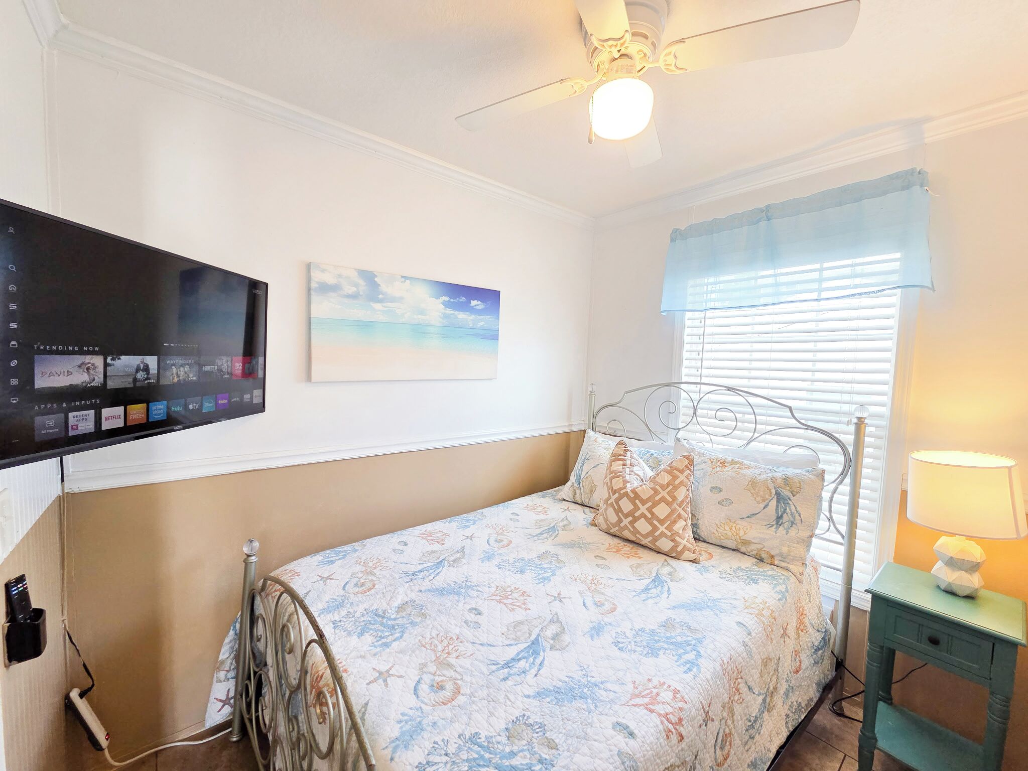 Een slaapkamer met een bed, een aan de muur bevestigde televisie, een plafondventilator, een raam met jaloezieën en een nachtkastje met een lamp.