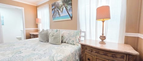 A bedroom with a bed, a dresser, a nightstand, a lamp, a painting, and a window with curtains.