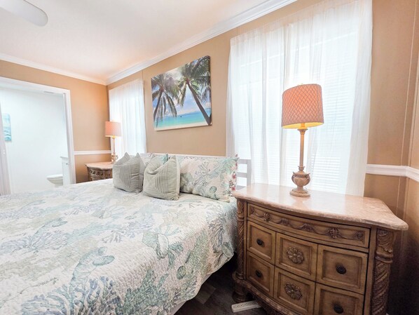 A bedroom with a bed, a dresser, a nightstand, a lamp, a painting, and a window with curtains.
