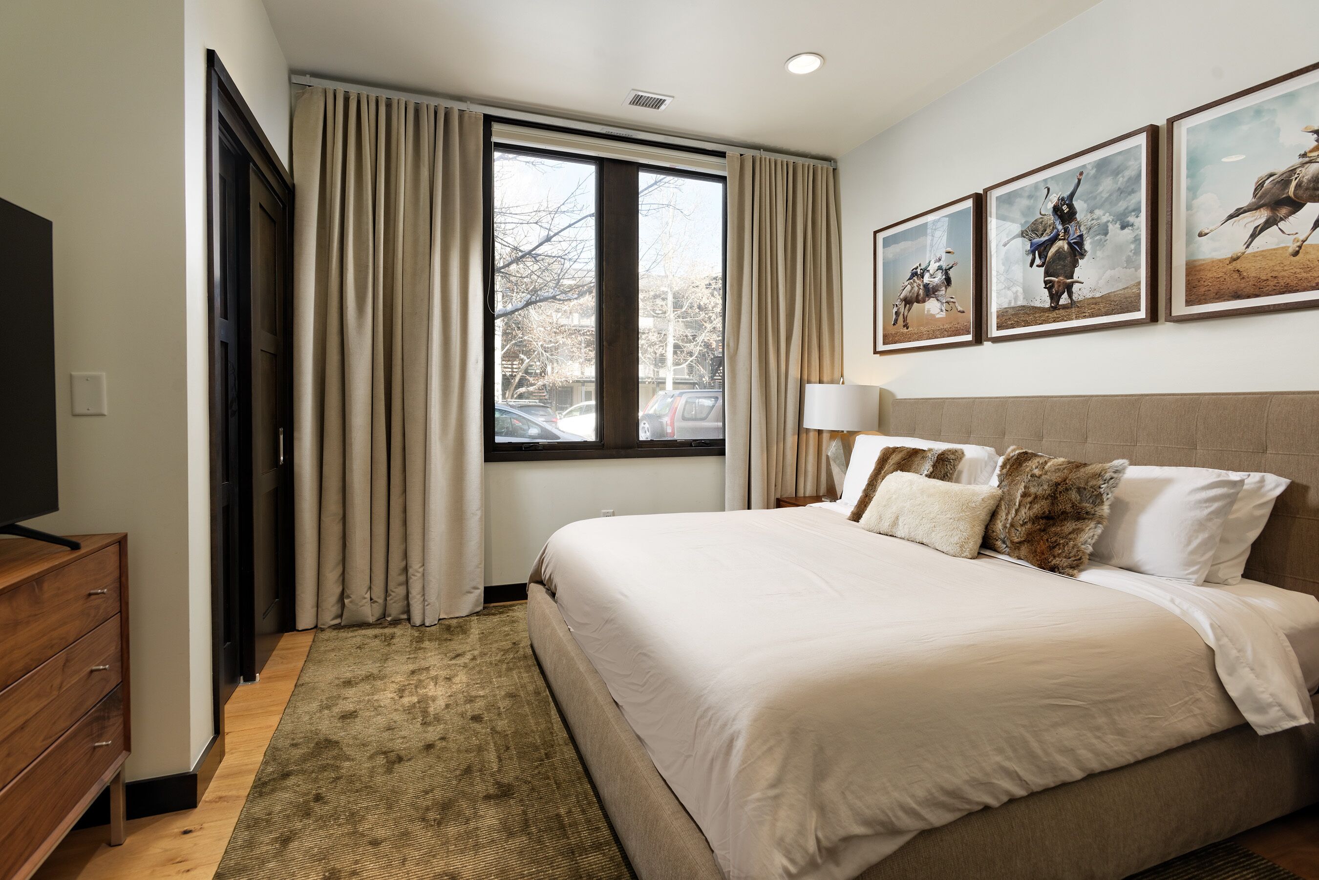 A bedroom with a large bed, a wooden dresser, a window with curtains, and three framed pictures on the wall.