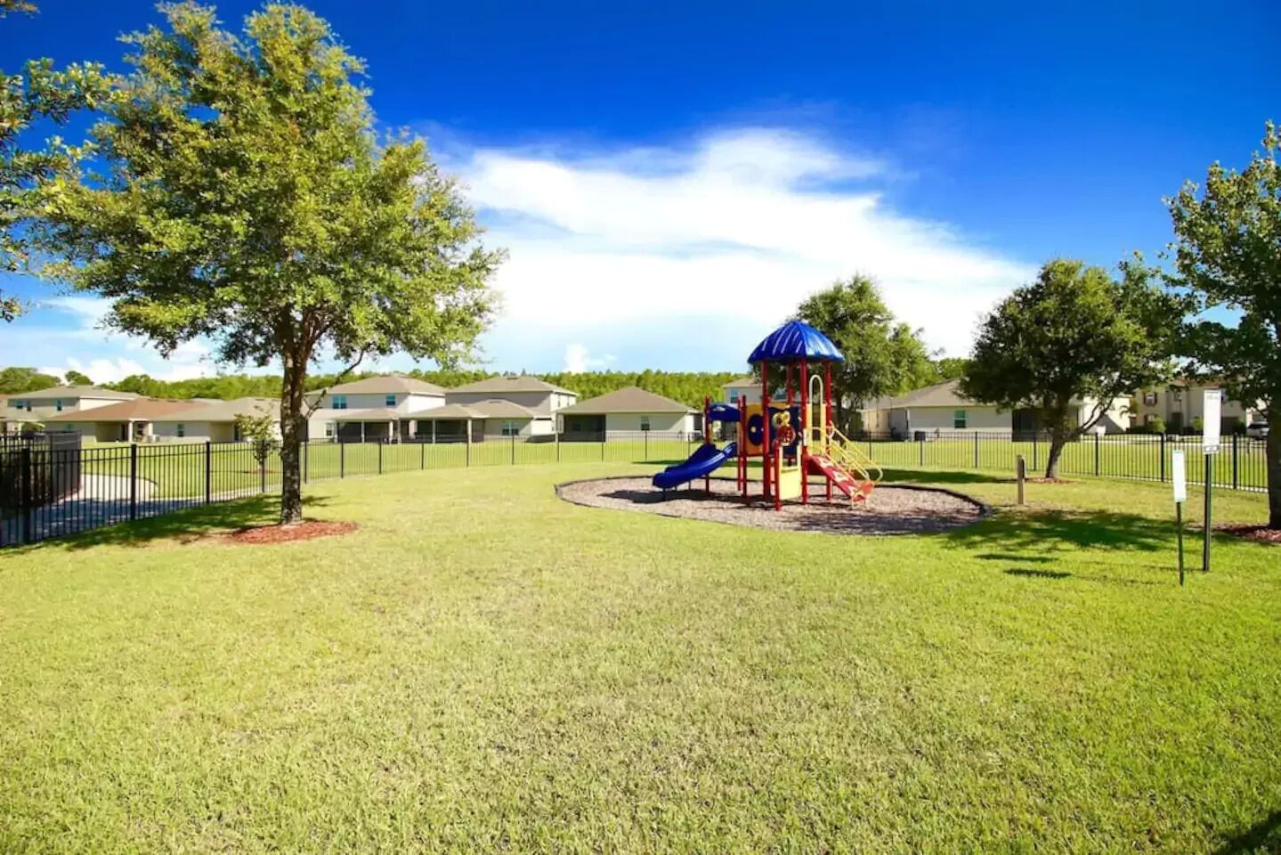 A playground with slides and a climbing structure, surrounded by a fenced area and trees.