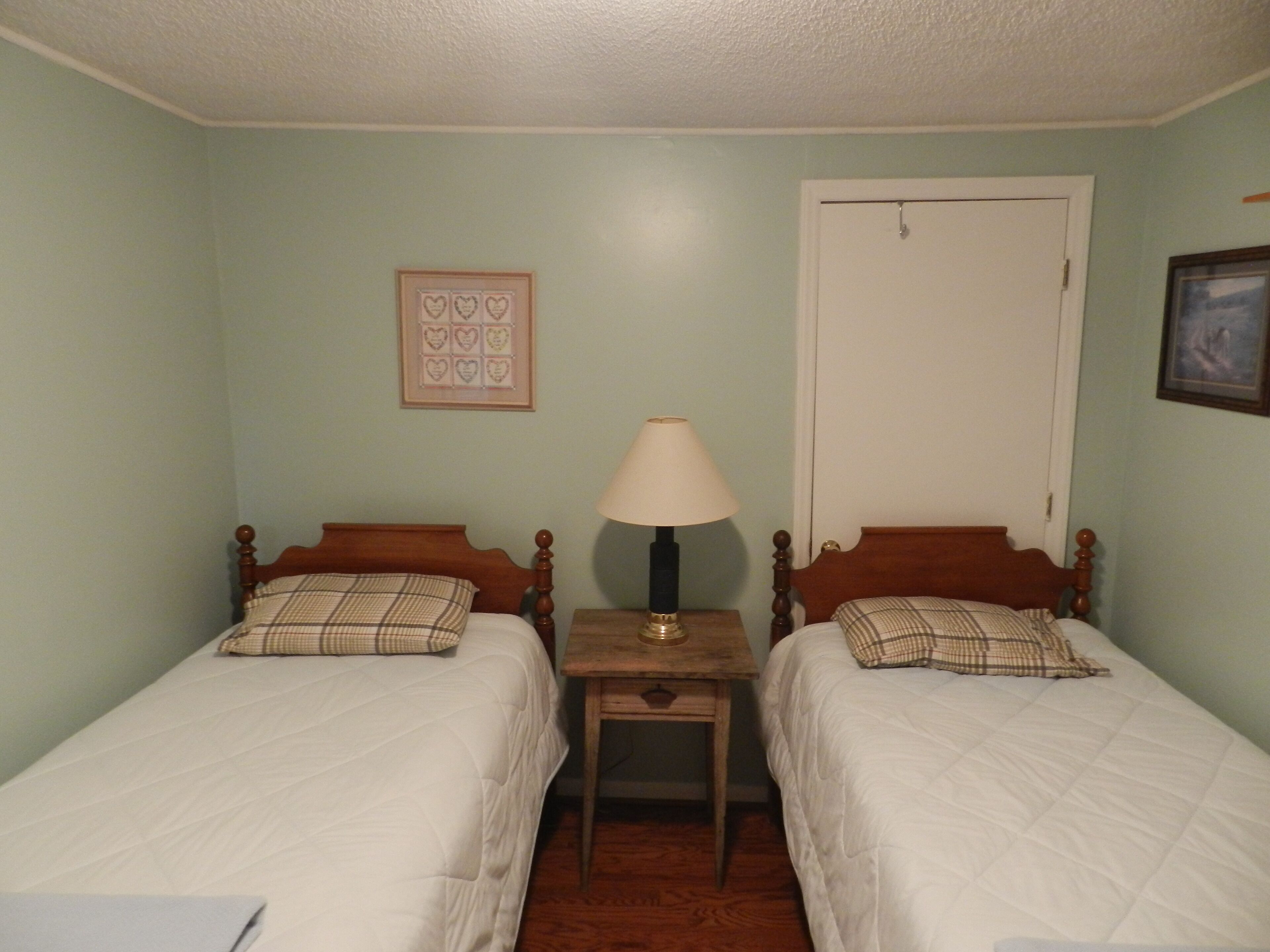 Quarto com duas camas, mesa de cabeceira com abajur, porta e quadro pendurado na parede.
