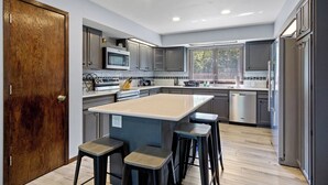 A modern kitchen with grey cabinets, a central island with stools, stainless steel appliances, and a window with blinds. - Family Adventure at Lake Wissota Lodge + Studio (Jim Falls)