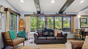 A living room with a sofa, a wooden chair, and a dining table. There are large windows with a view of trees. - Family Adventure at Lake Wissota Lodge + Studio (Jim Falls)