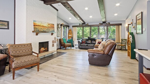 A spacious living room with a fireplace, wooden beams, and large windows offering a view of the outdoors. - Family Adventure at Lake Wissota Lodge + Studio (Jim Falls)