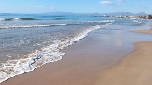 Une plage à l’eau d’un bleu limpide, avec de douces vagues et une chaîne de montagnes qui se dessine à l’horizon.