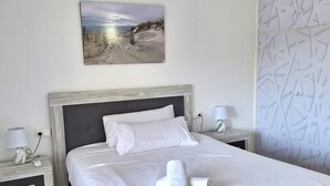 Un lit bien fait, avec du linge de lit blanc et une serviette enroulée, un tableau représentant une scène de plage accroché au mur et un panneau mural à motifs.