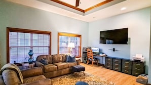 A spacious living room with a large sectional sofa, a coffee table, and a rug. There are two windows with blinds, a flat-screen TV mounted on the wall, and a desk with a chair.