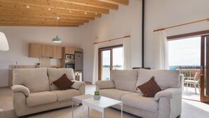 Un ampio soggiorno con due divani, un tavolino, travi in legno a soffitto, una cucina e grandi finestre con tende.
