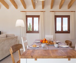 Un coin repas avec une table en bois mise pour le petit-déjeuner, deux fenêtres avec des rideaux et un canapé à l’arrière-plan.