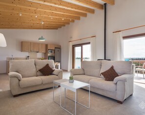 Un salon spacieux avec deux canapés, une petite table, des poutres apparentes en bois au plafond, une cuisine et de grandes fenêtres avec des rideaux.