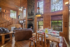 Interior de cabana de madeira com lareira, mesa de jantar posta para quatro pessoas, sofá e escada.