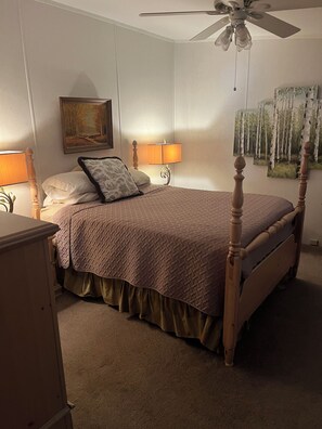 A bedroom with a four-poster bed, a bedside lamp, a wall-mounted painting, and a ceiling fan.