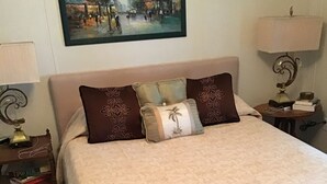 Una cama bien hecha con una manta color beige, dos almohadas decorativas y un cuadro enmarcado encima.