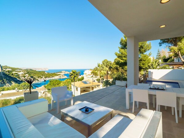 A terrace with white furniture, a pool, and a scenic view of the sea and hills.