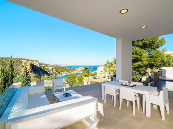 Un área de comedor al aire libre moderna con piscina, mobiliario blanco y una vista panorámica de la costa y los acantilados.