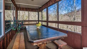 A covered outdoor dining area with a wooden table and benches, large windows, and a view of trees. - Pappy’s Hideaway (Gatlinburg)