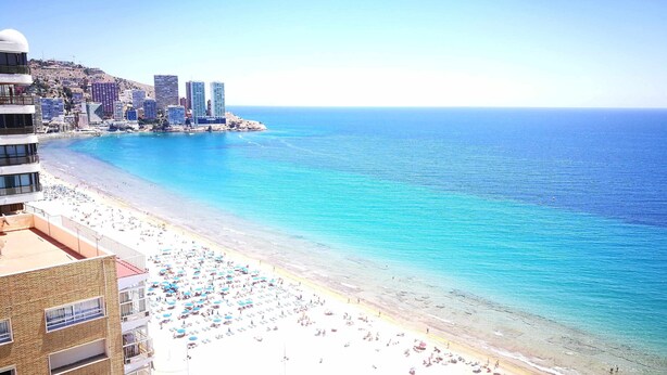 Ein Strand mit klarem, blauem Wasser, einem sandigen Ufer und einer Stadtkulisse im Hintergrund.