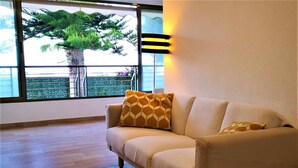 Un soggiorno con un divano beige, due cuscini decorati, un'ampia finestra con vista su una palma e un balcone, e un punto luce a parete.