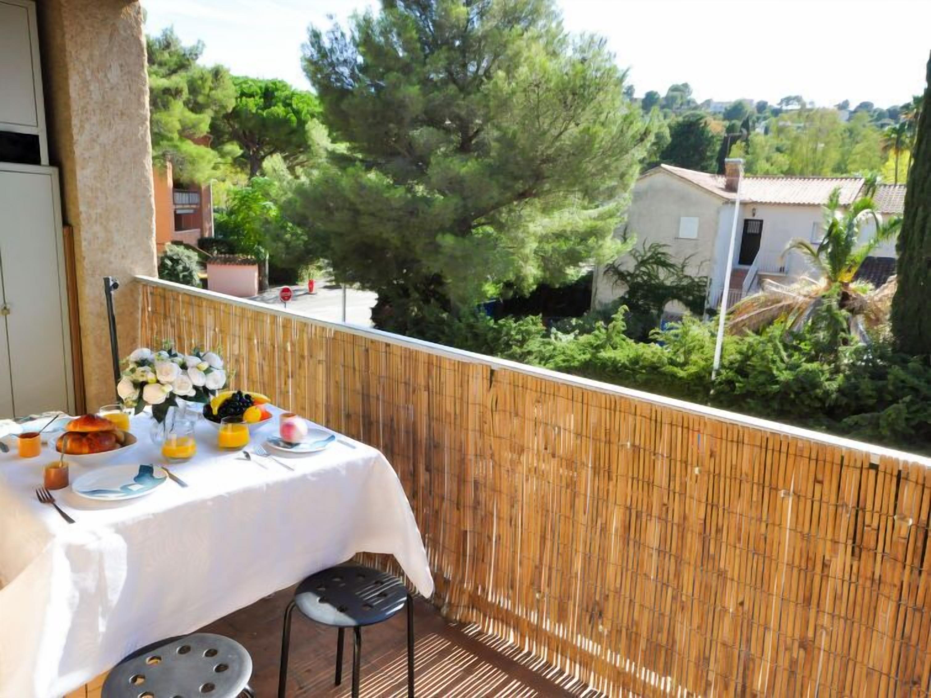 Un balcon avec une table dressée pour le petit-déjeuner, comprenant des croissants, des fruits et du jus, donnant sur un jardin luxuriant et des immeubles d’habitation.