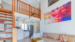 Un appartement moderne de style « loft », doté d’un escalier en bois, d’une cuisine, d’un espace de vie avec un canapé et des coussins, et d’une peinture abstraite aux couleurs vives accrochée au mur.