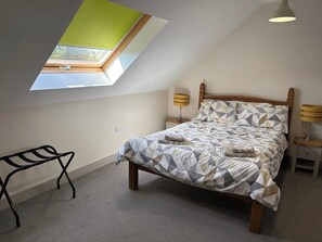 Um quarto com teto inclinado, um claraboia, uma cama com colcha estampada em padrão geométrico, duas luminárias de mesa e um pequeno banco de metal preto.