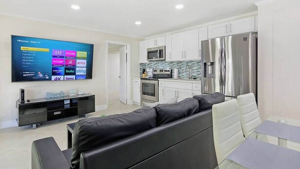 A modern living room with a black leather sofa, a flat-screen TV mounted on the wall, and a kitchen area with white cabinets and stainless steel appliances.