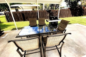 A glass-top patio table with four chairs, a barbecue grill, and a covered area with a wooden fence in the background. - Private Guest suite with a beautiful view of the new hard rock hotel and casino (Fort Lauderdale)