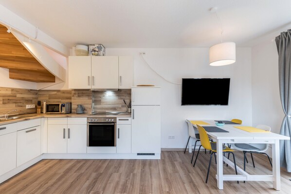 Uma cozinha moderna com armários brancos, micro-ondas e fogão. Há uma mesa de jantar com cadeiras e uma TV de tela plana fixada na parede.