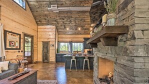 A spacious interior with a wooden ceiling, a stone fireplace, a kitchen area, and a seating area with a rug.