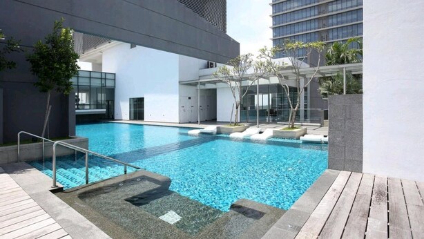 A modern outdoor swimming pool with a wooden deck, surrounded by a building with large windows and a covered seating area. - GT Home Cascades Residency (Petaling Jaya)