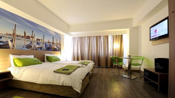 A hotel room with a bed, a painting of a harbor scene, a television, and a desk with a chair. - Ibis Styles Skopje (Skopje)