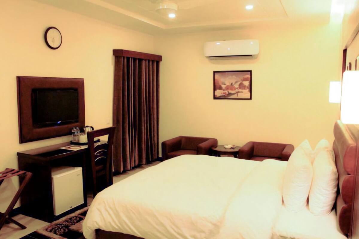 A hotel room with a bed, a desk with a television, a chair, a small table, and an air conditioning unit.