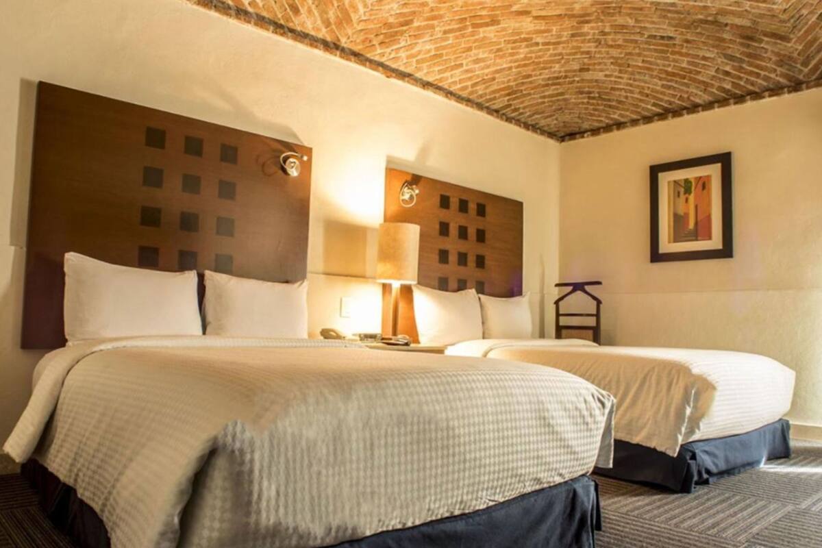 A hotel room with two beds, a brick ceiling, a nightstand with a lamp, and a framed picture on the wall.