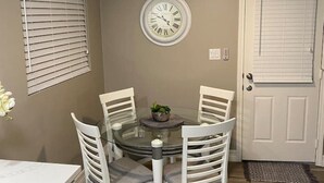 Uma sala de jantar com uma mesa de vidro redonda, quatro cadeiras brancas, um ventilador de teto e um relógio de parede.