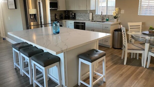Cozinha moderna com ilha grande, armários brancos, eletrodomésticos de aço inoxidável e área de jantar com mesa de vidro e cadeiras brancas.