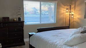 Quarto com uma cama grande, cômoda, mesa de cabeceira e janela com persianas.