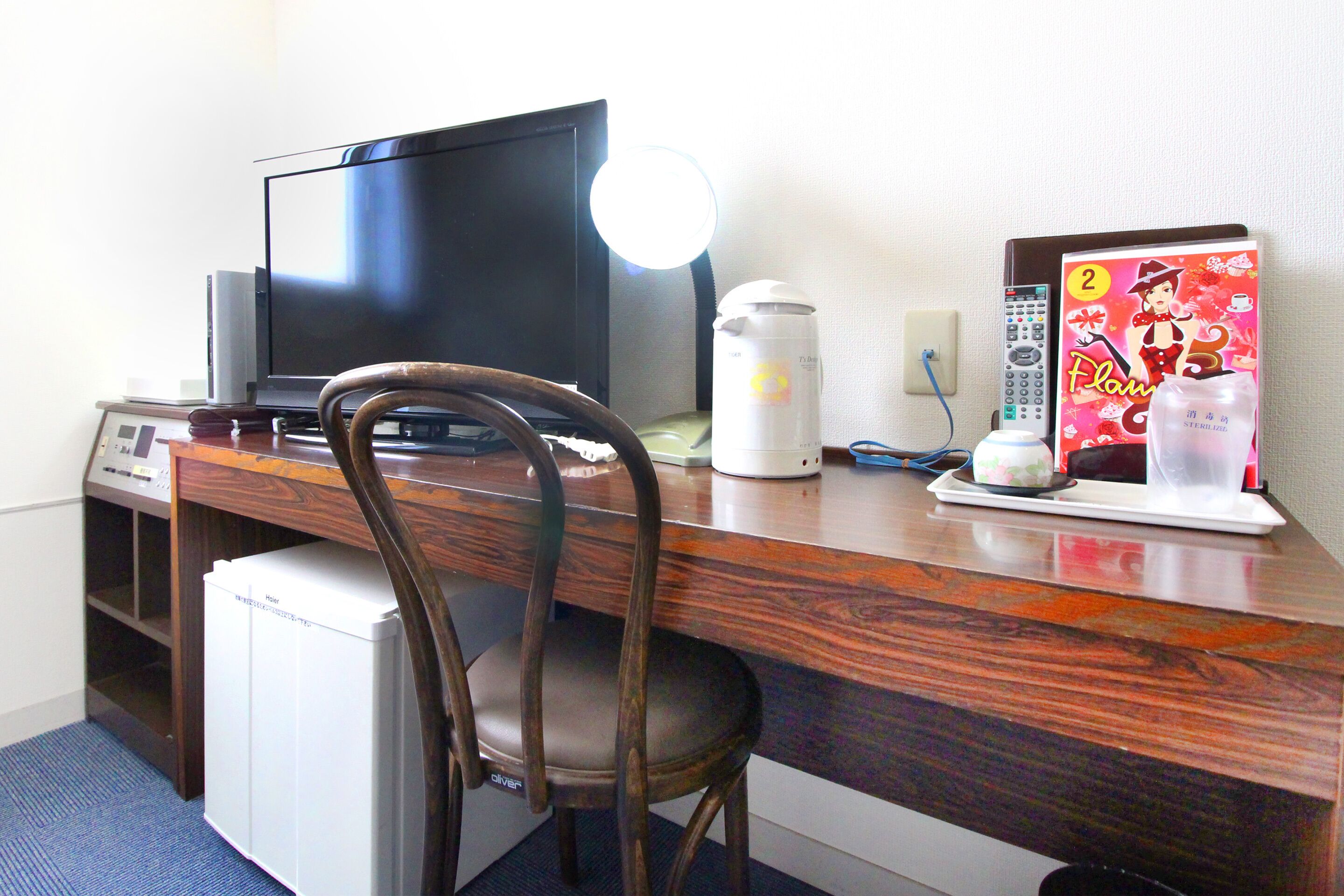 Habitación de hotel con escritorio de madera, televisión, refrigerador pequeño, una silla, lámpara, control remoto y un adorno en la pared.